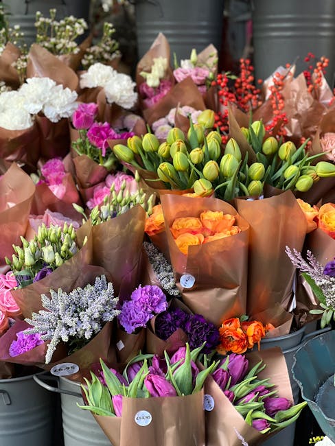 A diverse display of fresh flowers arranged in brown kraft paper wrapping, including clusters of white hydrangeas, vibrant pink carnations, orange roses, purple statice, and green tulips with closed b