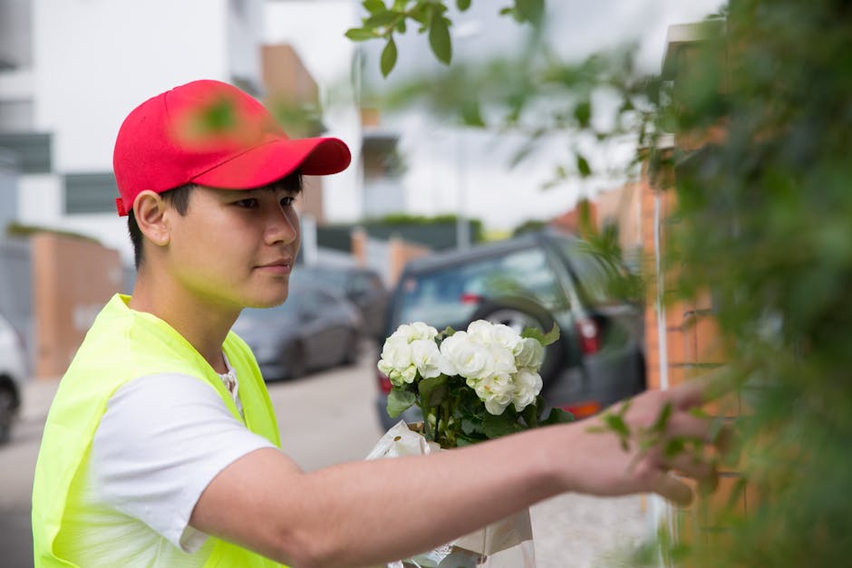 A young delivery person wearing a red cap, white t-shirt, and high-visibility yellow vest is reaching out to place a bouquet of fresh white hydrangeas wrapped in brown paper against a brick wall. The
